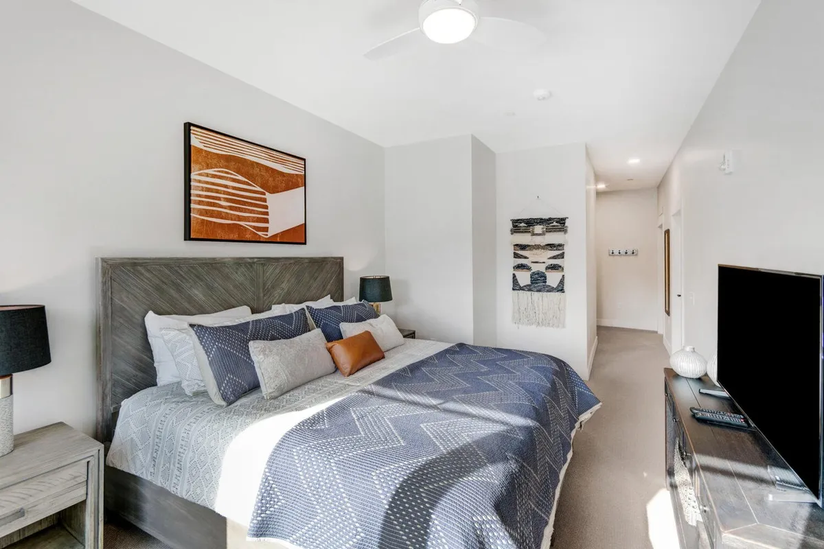 A serene bedroom featuring a bed with plush, patterned bedding, abstract artwork above the headboard, two nightstands with stylish lamps, and a television on the opposite wall. Natural light filters in, creating a tranquil and cozy atmosphere perfect for rest.