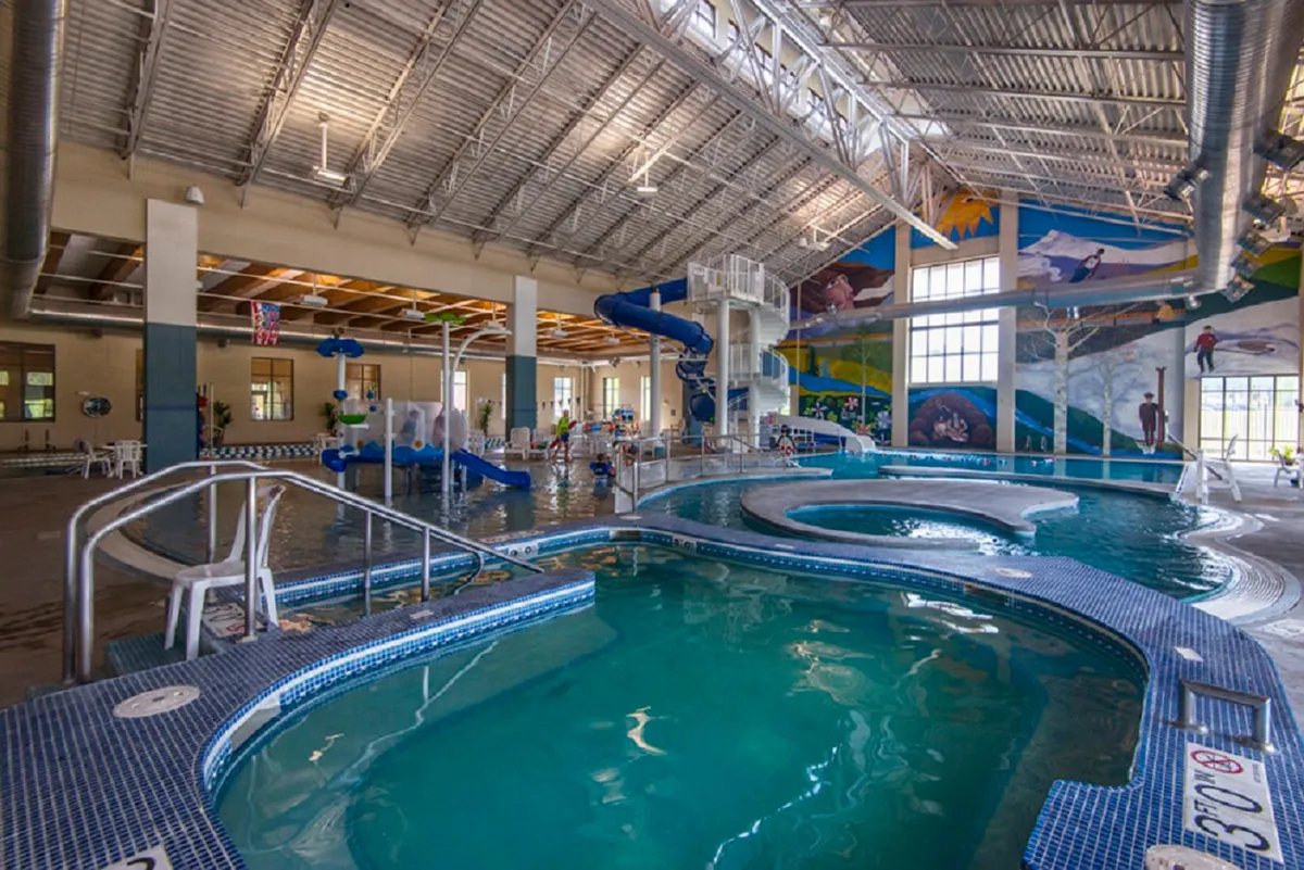 An energetic indoor pool scene at the Grand Park Recreation Center, featuring colorful water slides, a swimming pool, children playing, and cheerful murals. The setting is bright and full of family activity.