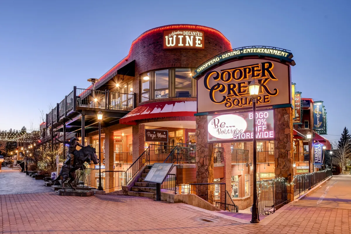 A lively outdoor plaza with the Cooper Creek Square sign, a wine shop, outdoor seating, and people gathering for shopping, dining, or entertainment. The cheerful mood reflects the convenience of having restaurants, shops, and events just steps away.