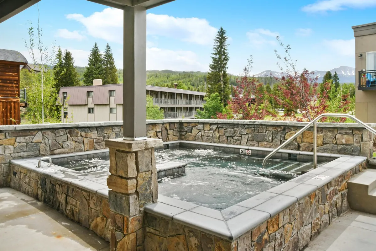 This image showcases a large hot tub set against a backdrop of serene nature and mountains. The hot tub area appears expansive, capable of hosting a group, with tranquil surroundings enhancing the sense of luxury and relaxation. The scene is peaceful, inviting, and embodies the essence of resort-style comfort.