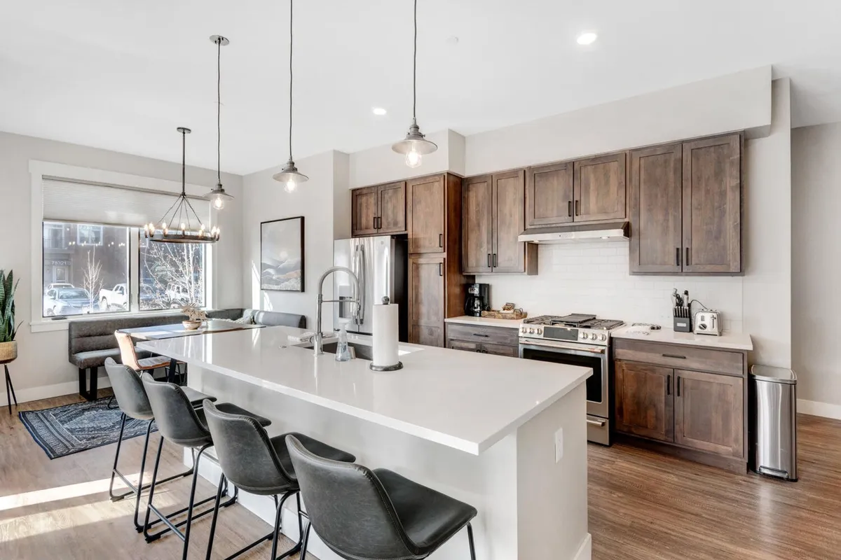 A modern kitchen scene with a spacious island countertop, stainless steel appliances, contemporary light fixtures, and a view toward the dining area. The setting is equipped for both gourmet cooking and casual snacks, with ample space for multiple people to gather and interact.