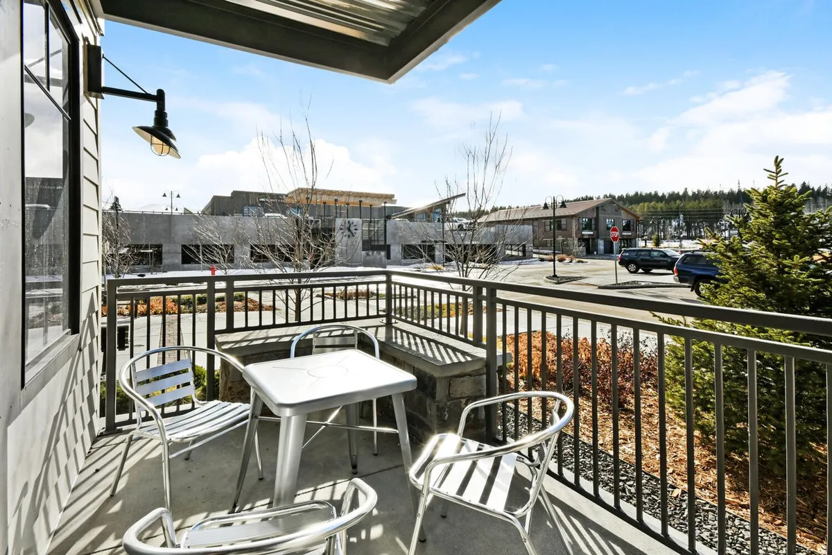 An inviting patio table with metal chairs is nestled amidst lush greenery. The scene is serene and casual, ideal for morning coffee, reading, or socializing, and reflects seasonal changes in foliage.