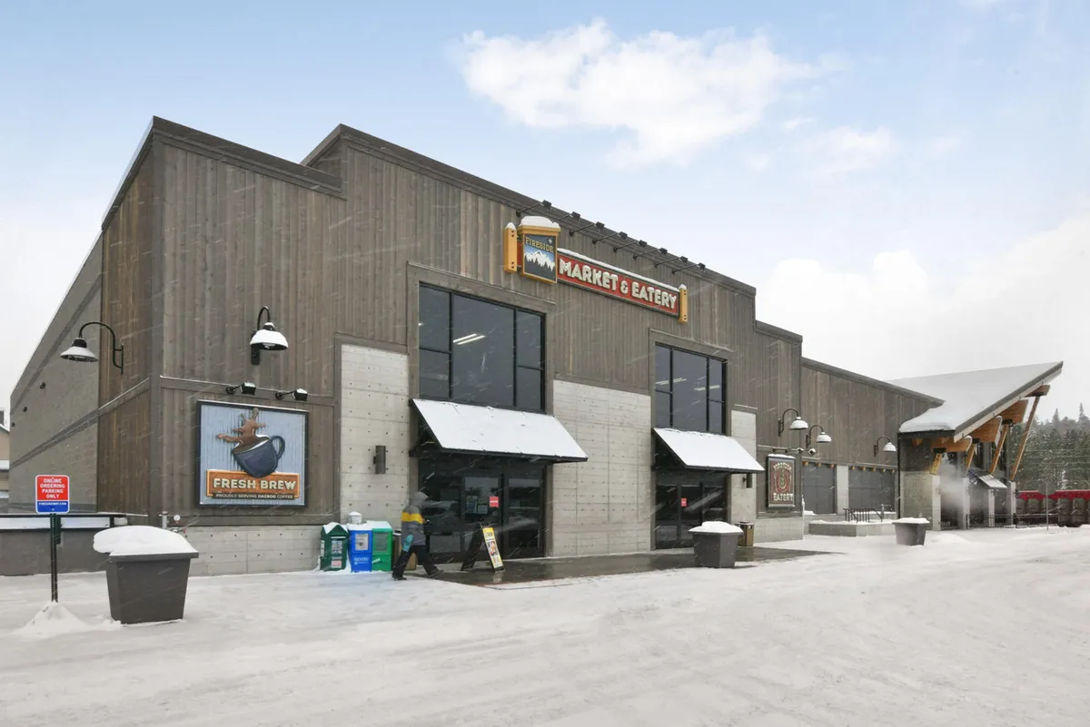 A cozy exterior view of a local market entrance, featuring welcoming signage, doors framed with rustic wood, and an outdoor seating area ideal for socializing or enjoying a warm beverage while taking in the mountain town atmosphere.