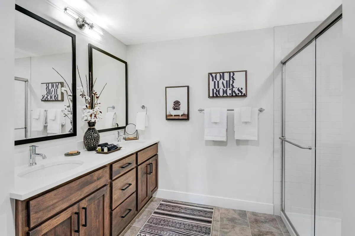 A modern bathroom featuring dual mirrors over a wooden vanity, a tasteful decorative vase, framed wall art, and fresh towels. Ample morning light enhances the calm, spa-like atmosphere for restful routines.