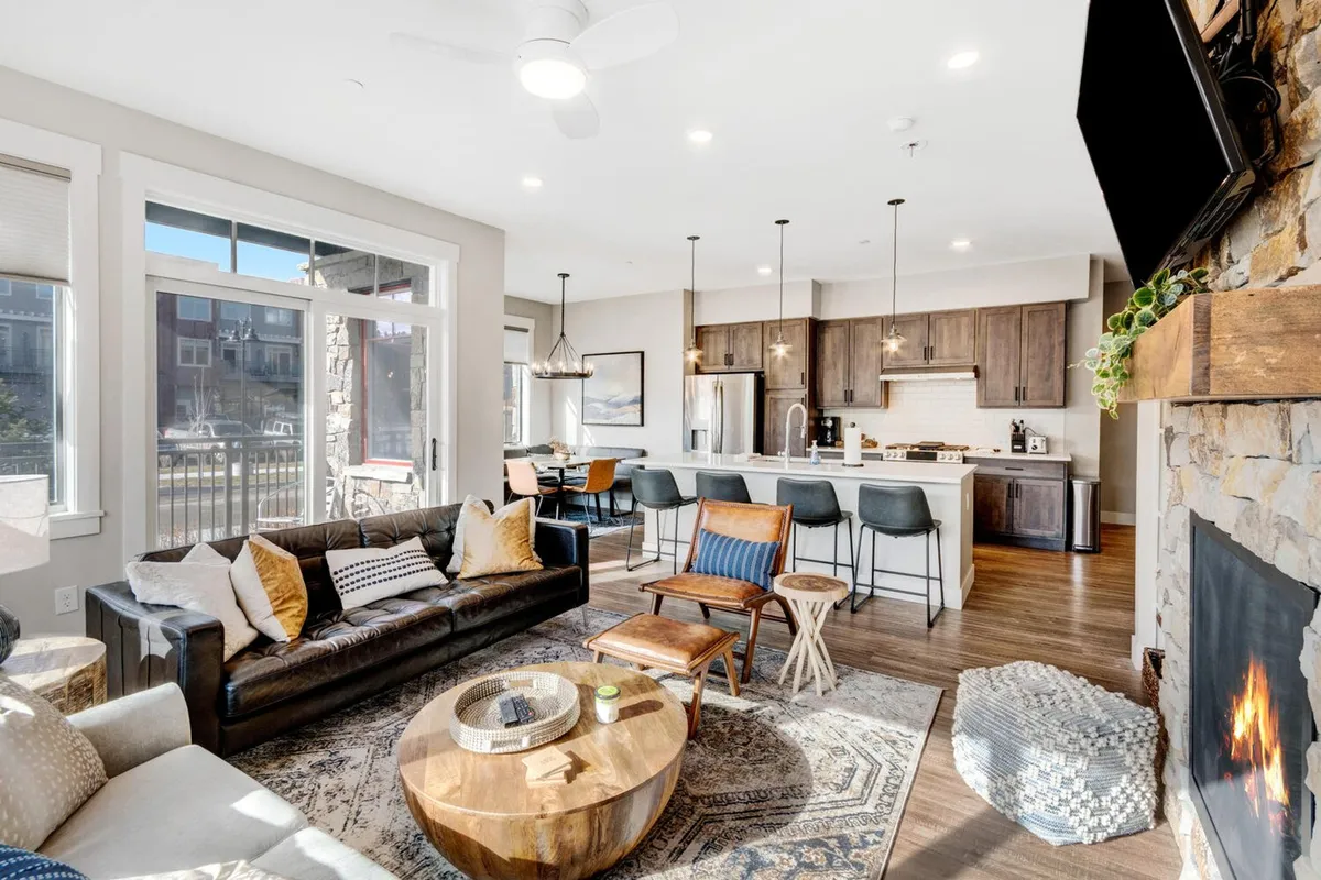 The image showcases a cozy and stylish open-plan living space with a plush sofa, rustic wooden coffee table, and a glowing fireplace as the centerpiece. The dining area is set for a gathering, and large windows hint at beautiful mountain views, creating a warm and inviting atmosphere perfect for relaxation and quality time.