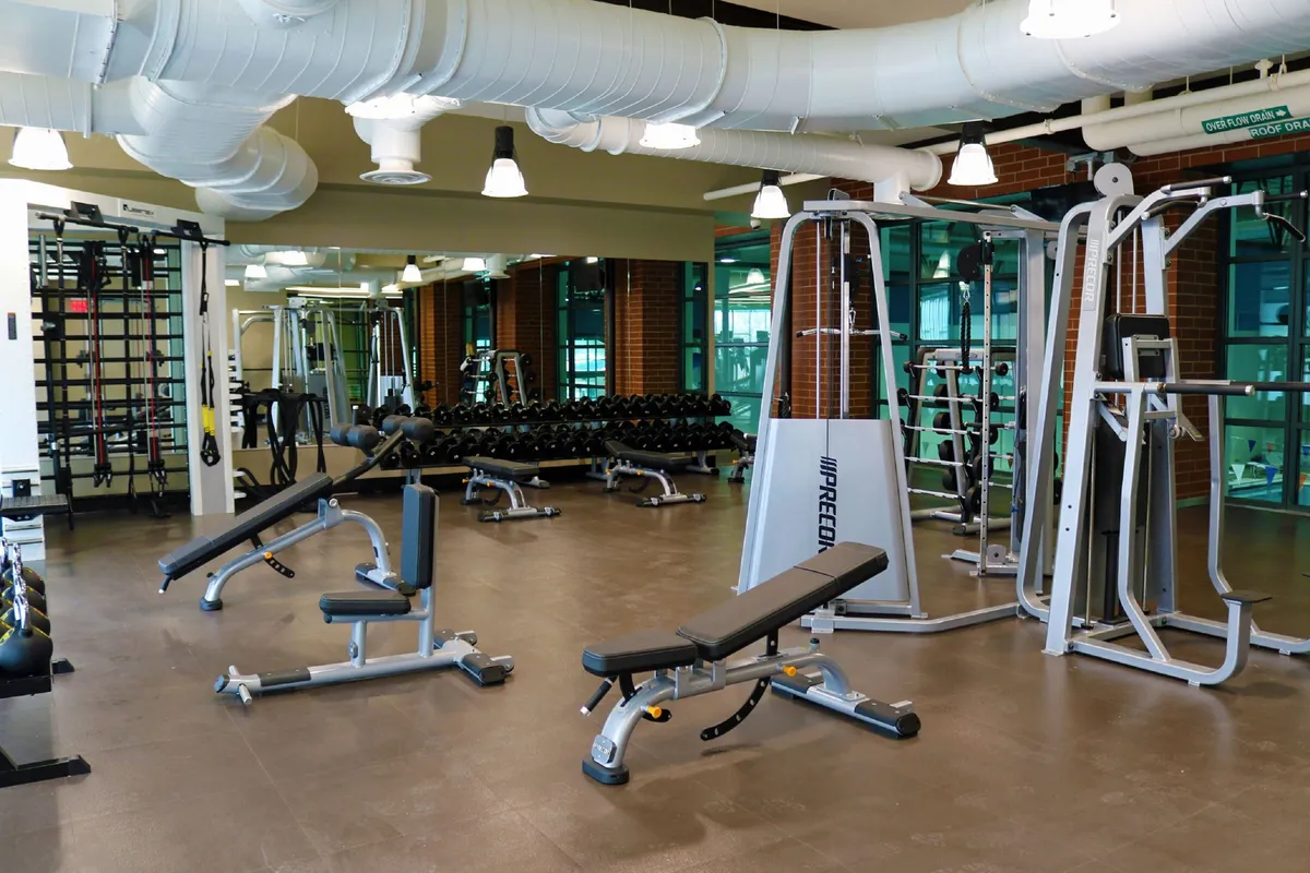 A well-equipped fitness room featuring weight benches, dumbbells, exercise machines, and mirrors. The space is designed for both individual training and group fitness activities, creating a motivational and energetic environment.