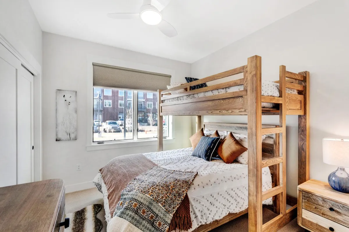 A welcoming second bedroom with a full-over-queen bunk bed, dressed with decorative pillows and set beside a bright window. The cozy nightstand with a lamp creates a warm, family-friendly vibe ideal for flexible sleeping arrangements.