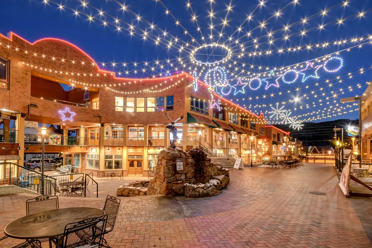 An inviting town center scene featuring twinkling overhead lights, decorative star shapes, a central stone fountain, and brick-paved walkways. People can be seen dining al fresco, browsing at boutiques, and enjoying a lively, festive atmosphere beneath the illuminated sky.