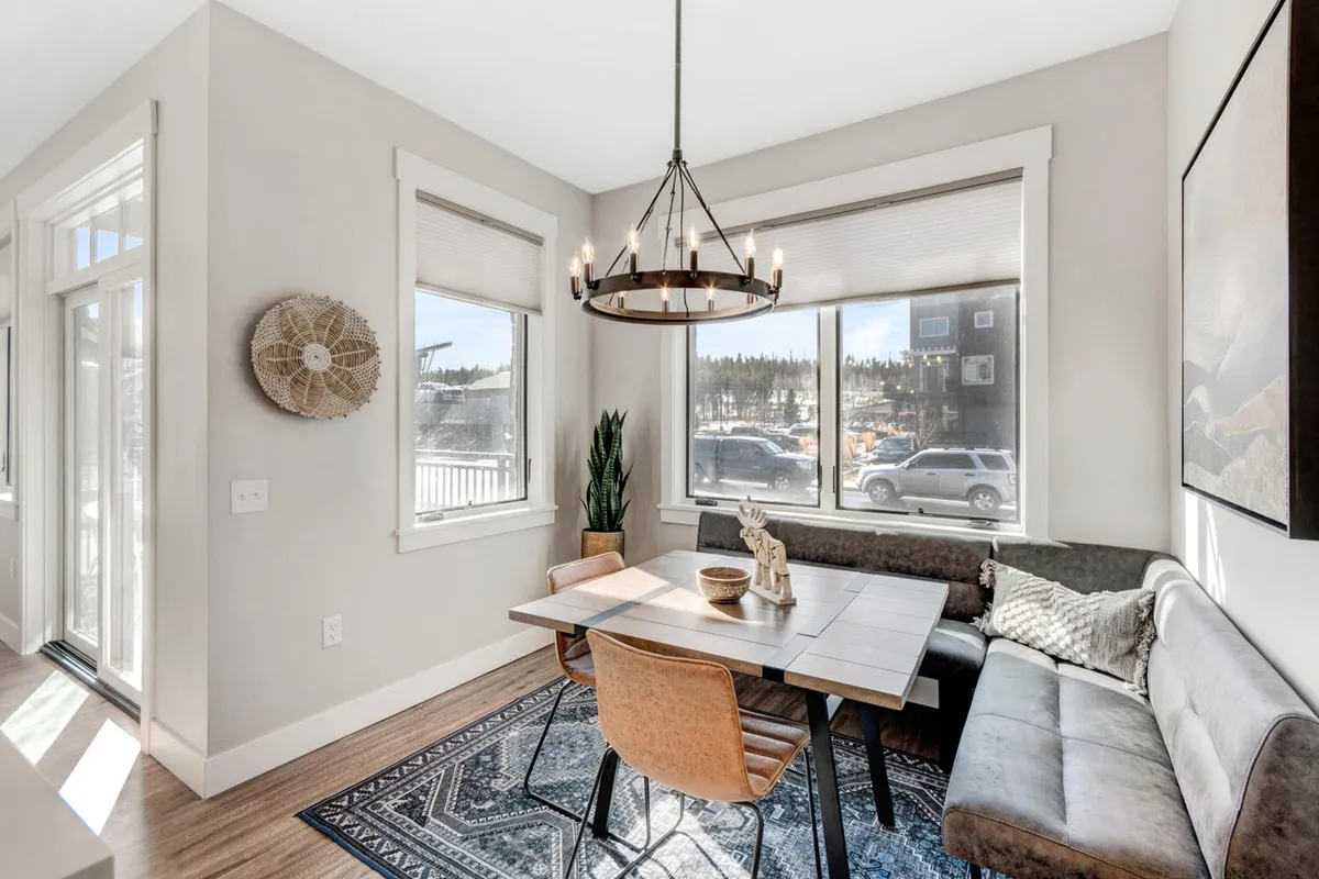 A close-up of the dining area featuring a stylish table, sofa, chandelier, and large windows that flood the room with natural light. The cozy arrangement and decorative touches create a welcoming setting for meals, board games, or morning coffee with views.